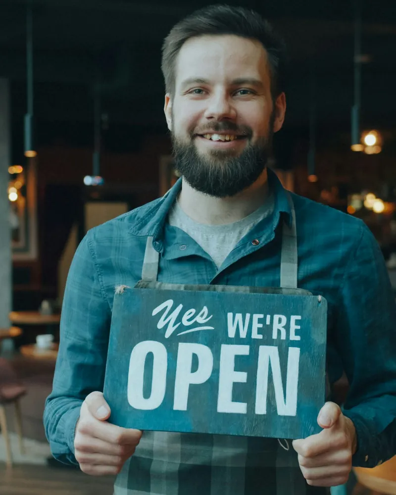A café owner in an apron smiling and holding a &ldquo;Yes, we&rsquo;re open&rdquo; sign